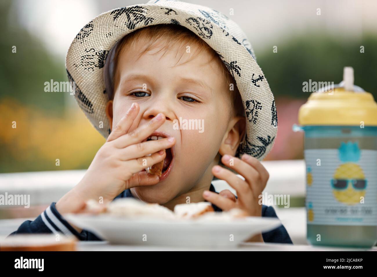 Close up image of Caucasian baby boy with blonde hair and hat opening mouth going to bite crispy bread, having joyful facial expression. Childhood, fo Stock Photo