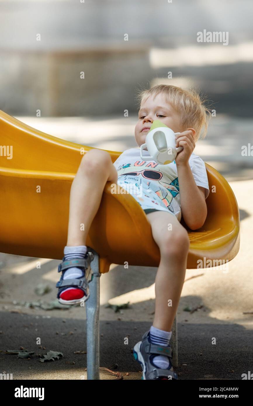 The little boy slips into a yellow plastic slide, his hair becomes eclectic, child sits down comfortably to calm and drink. Stock Photo