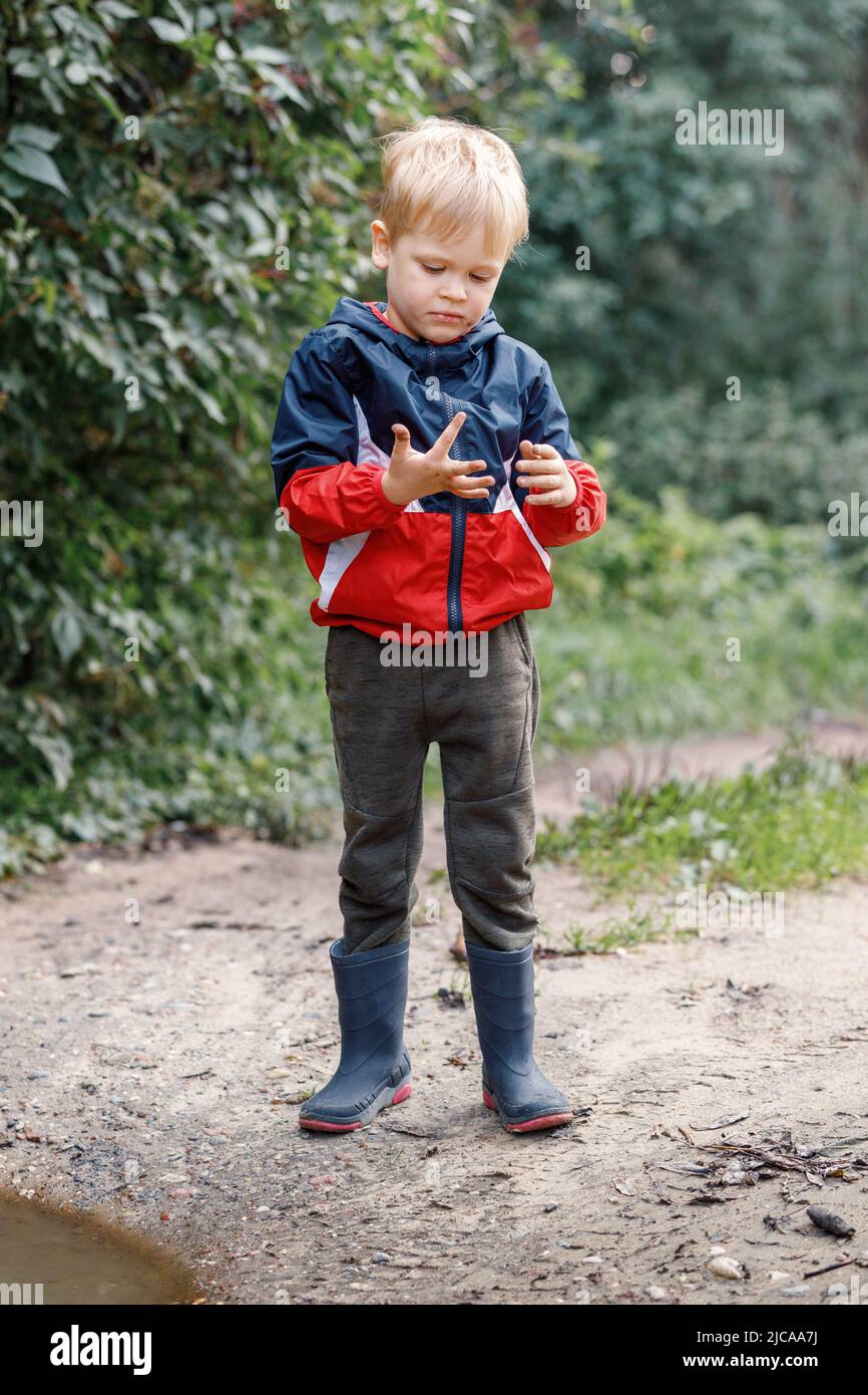 A little boy explores the mud near a puddle during a walk in nature, he looks at his smeared hands. Stock Photo