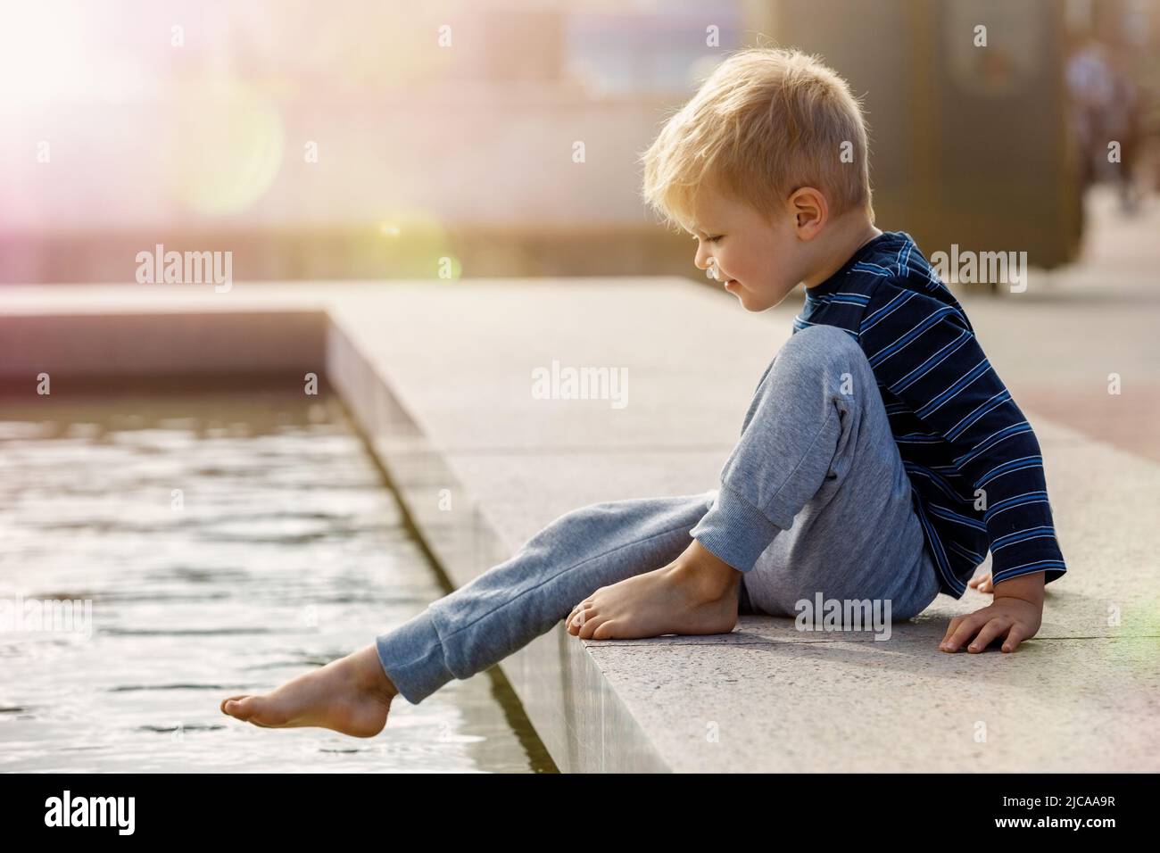 A cute little boy, barefoot, wants to climb into the water at the city fountain during the summer. Stock Photo