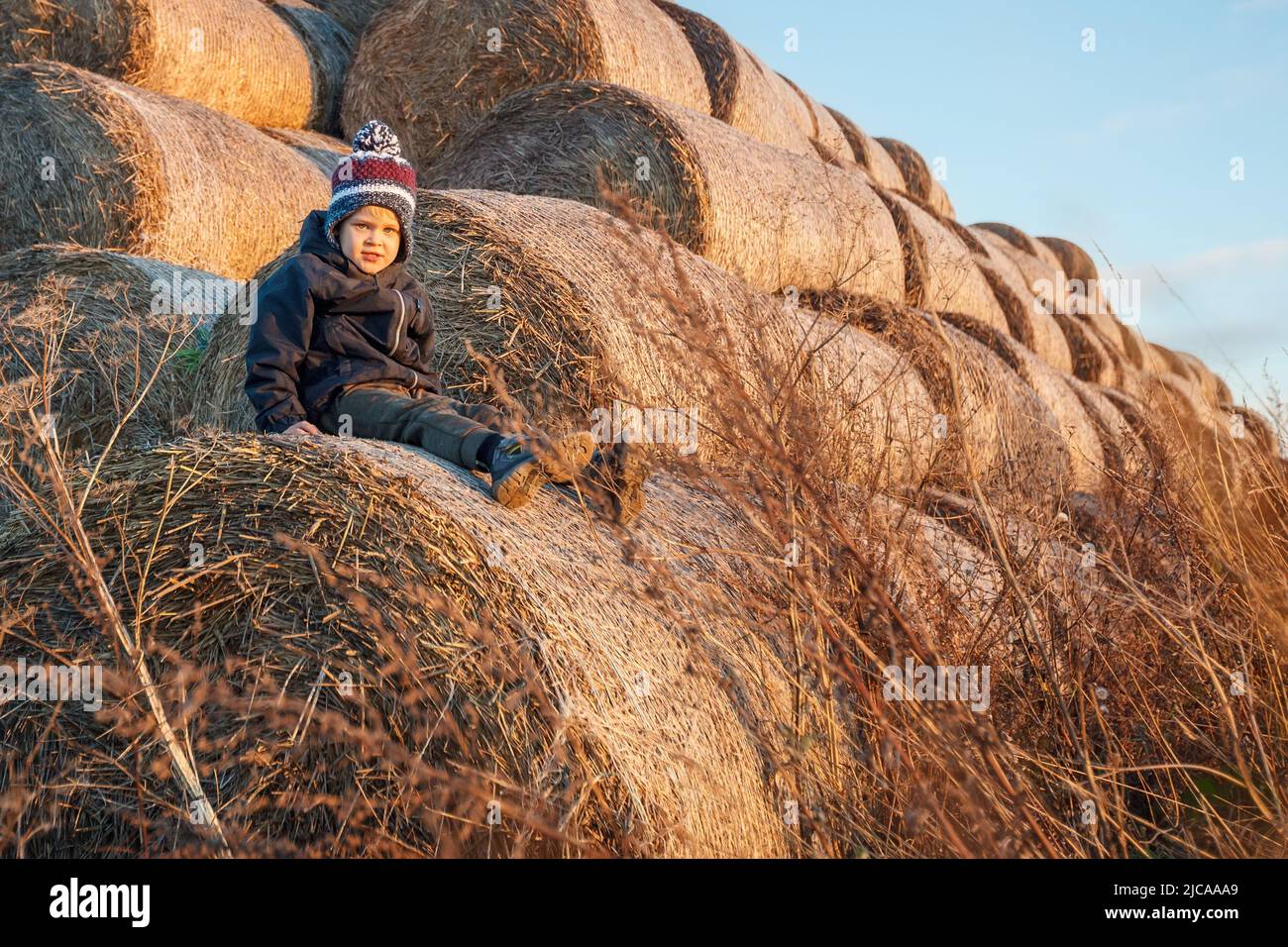 A cute little boy in autumn clothes and a knitted hat poses against a background of golden hay bales. Horizontal photo. Stock Photo