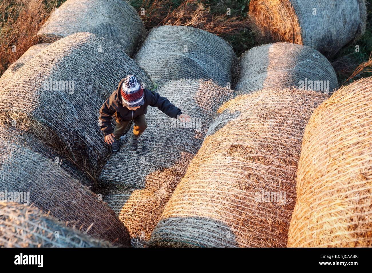 The little boy, in the autumn evening, in the sunset light, climbs up the hay rolls. Photographed from above. Stock Photo