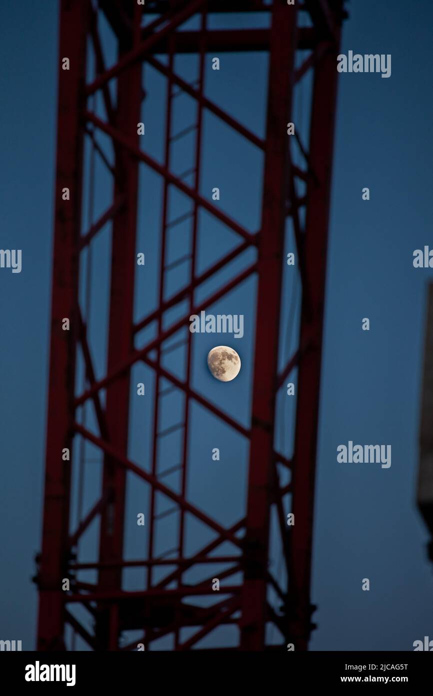 The moon is almost full and is visible through the red structure of a crane. Stock Photo