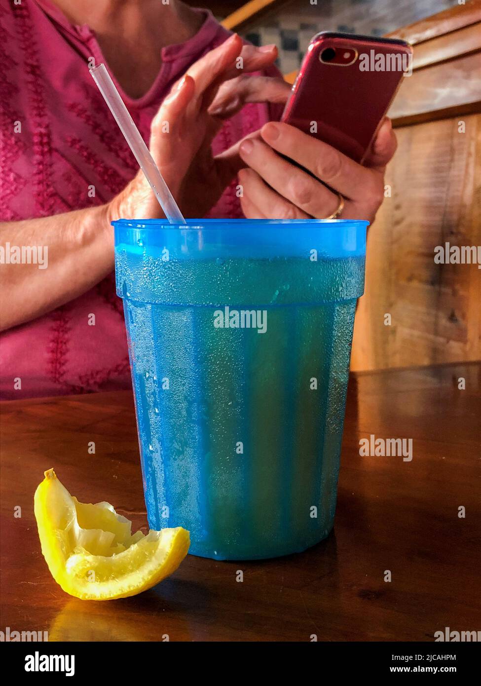 Cold beverage in blue plastic glass with condensation and a straw and a squeezed lemon wedge on the table, and a womans hands using using a mobile cel Stock Photo