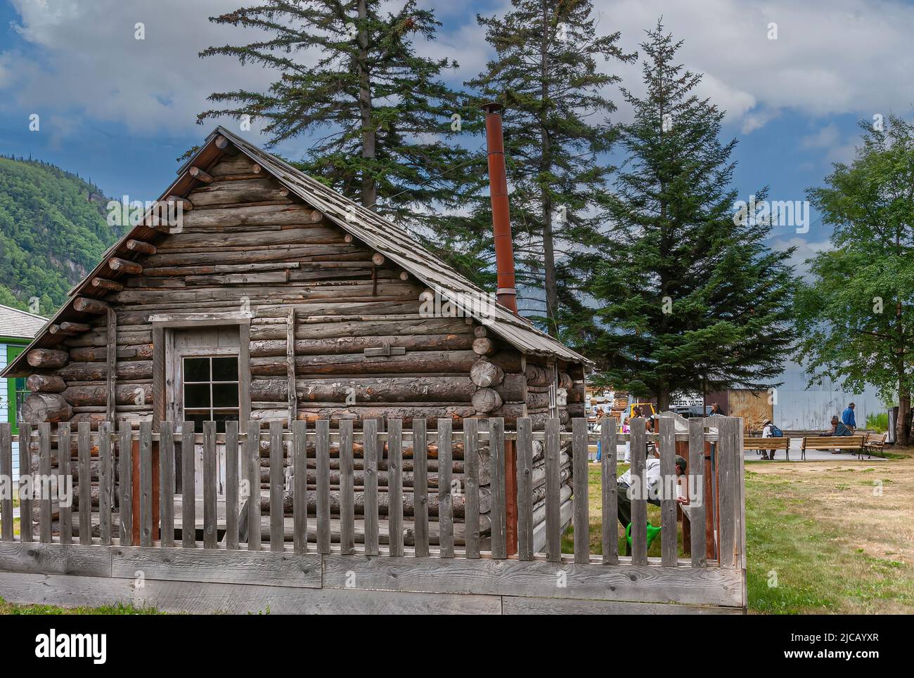 Skagway, Alaska, USA - July 20, 2011: Gray wooden small building is Historic Moore Homestead under blue cloudscape. Green foliage around and on mounta Stock Photo