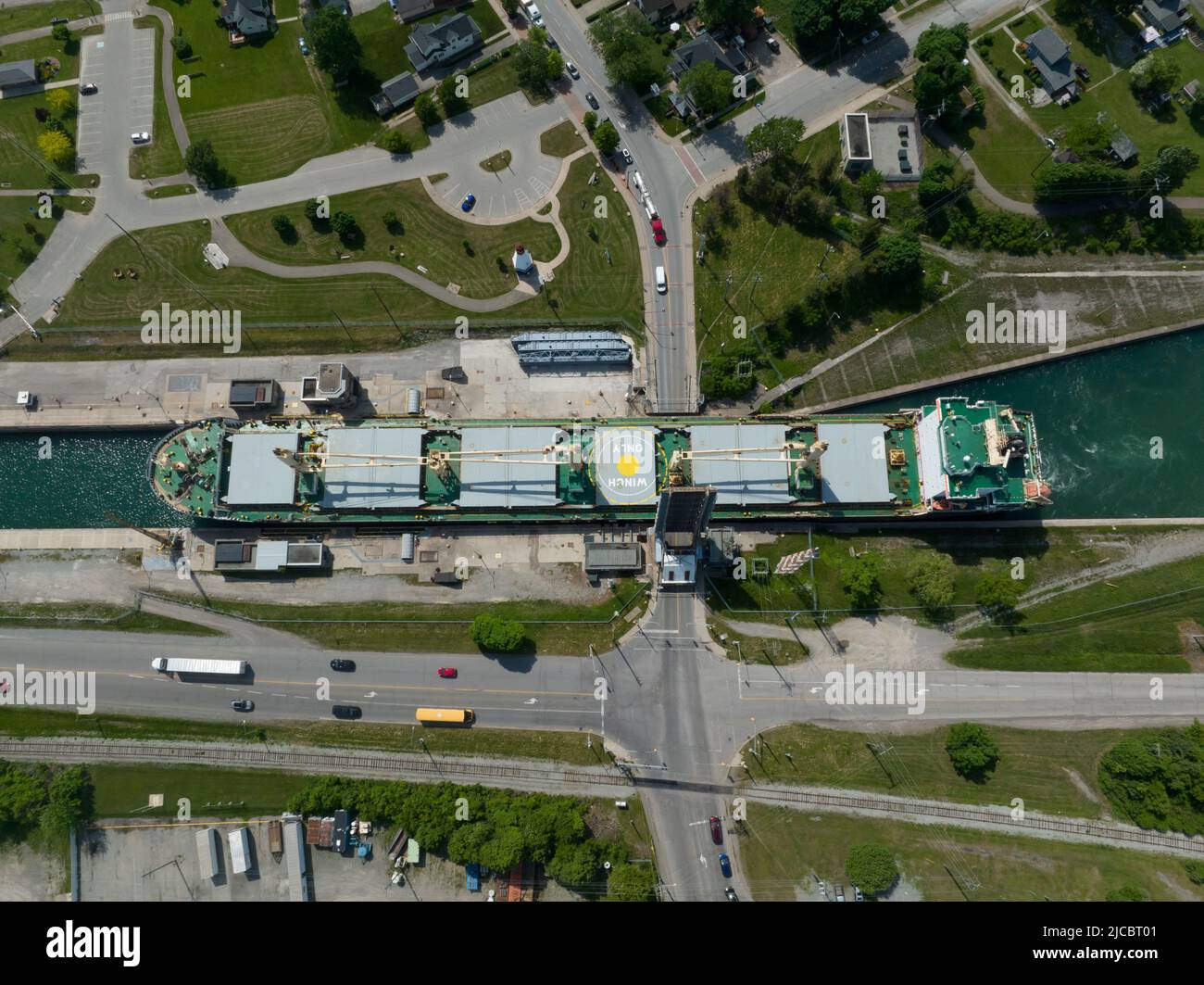 A direct overhead, top-down aerial view of a bulk carrier cargo ship passing through a tight urban canal at a drawbridge crossing during the day. Stock Photo