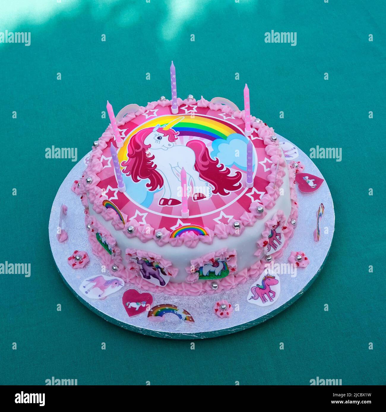 Homemade six year old girls birthday cake with unicorn theme decorated with rainbows stars hearts candles pink and white icing on silver cake board UK Stock Photo
