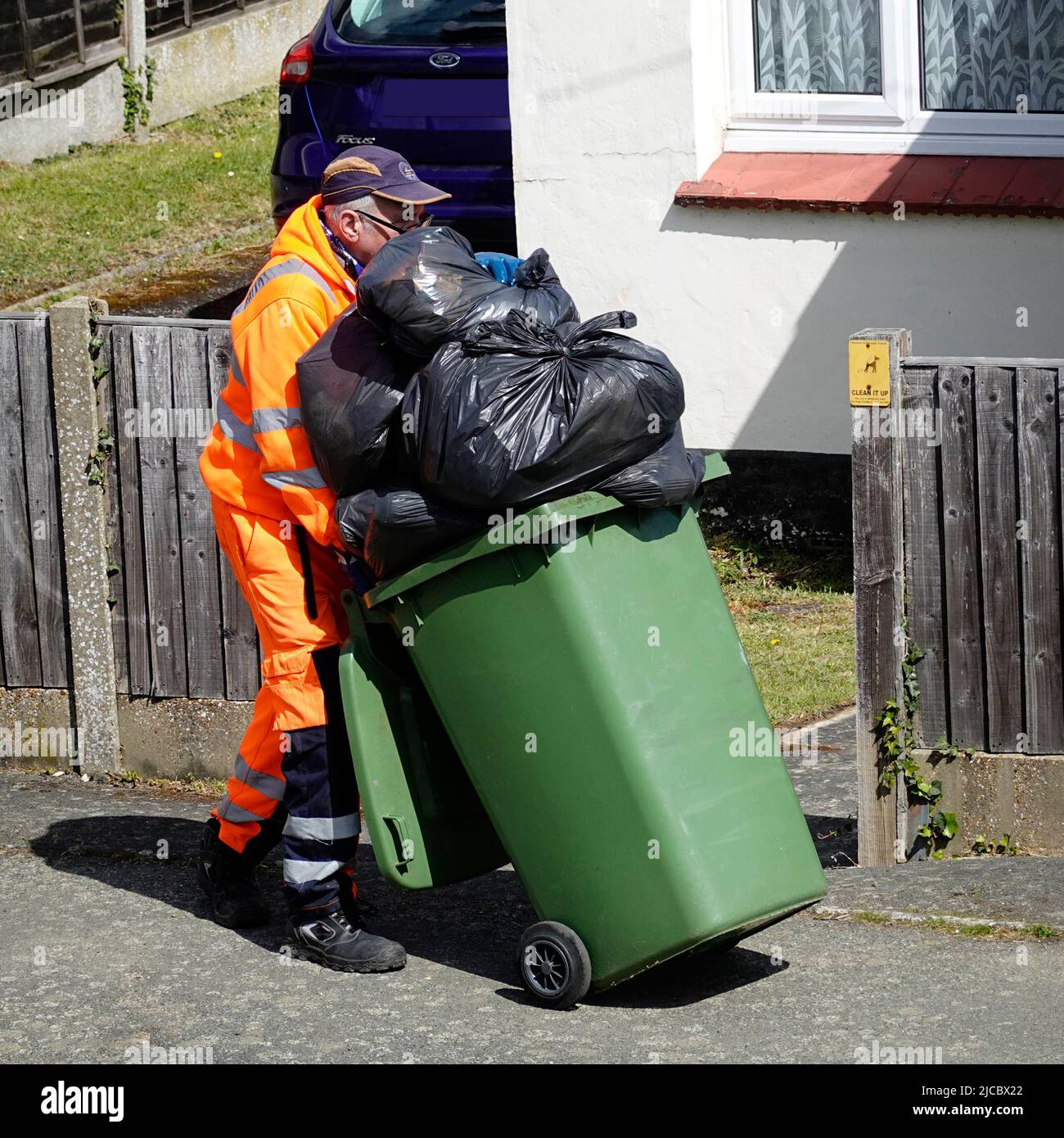 Waste management refuge collector dustman high visibility work clothing pushing overflowing wheelie bin full of black rubbish sacks Essex England UK Stock Photo