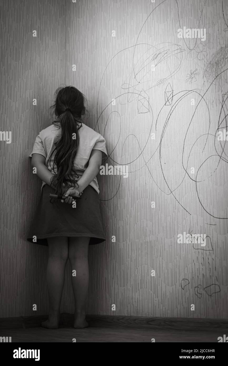 Black and white portrait of little bad girl holding marker behind back standing punished in corner for drawn wallpaper.  Stock Photo