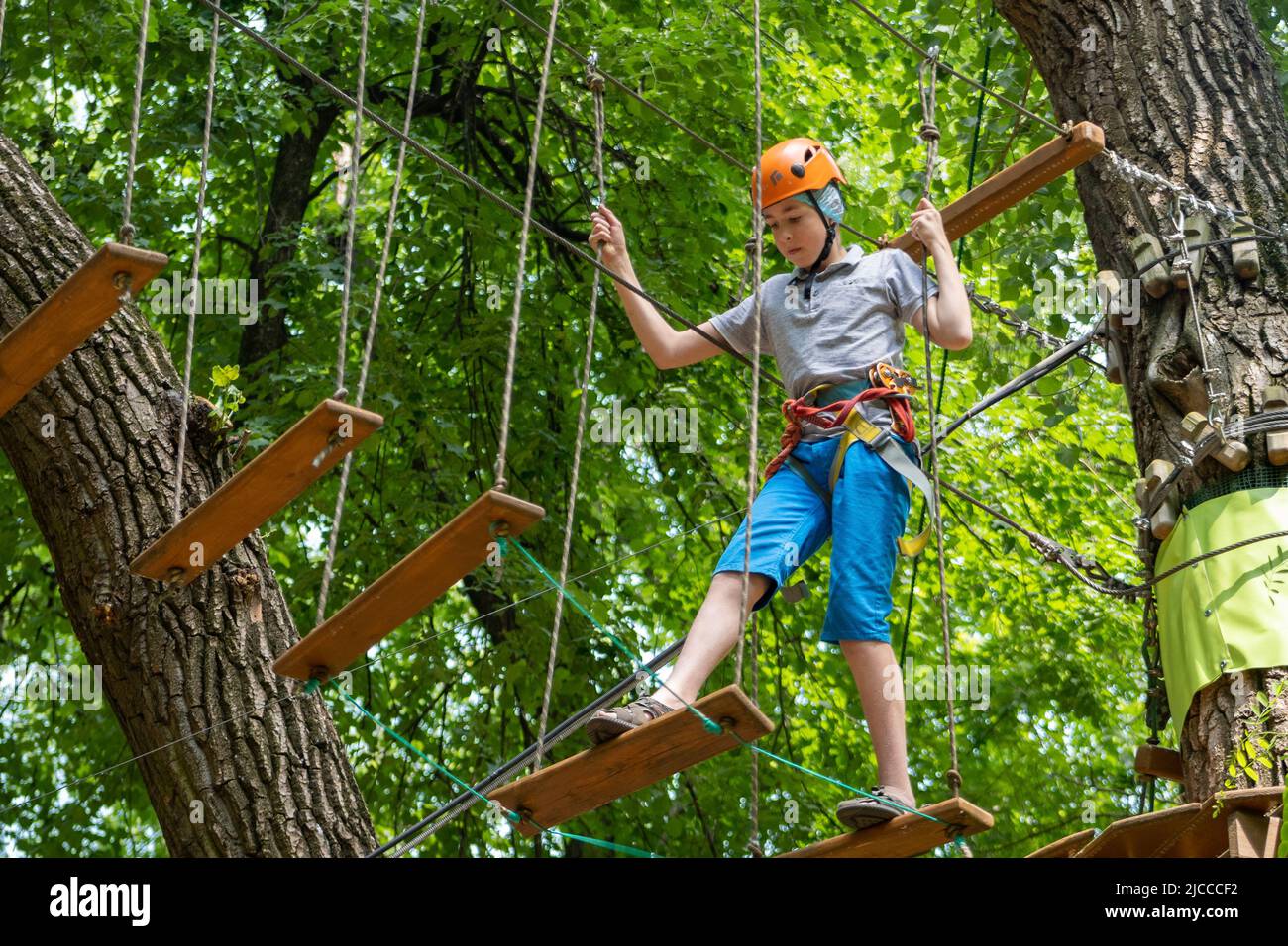 Rope park. A boy teenager in a helmet walks on suspended rope ladders. Carabiners and safety straps. Safety. Summer activity. Sport. Children's playground in nature in the forest. Stock Photo