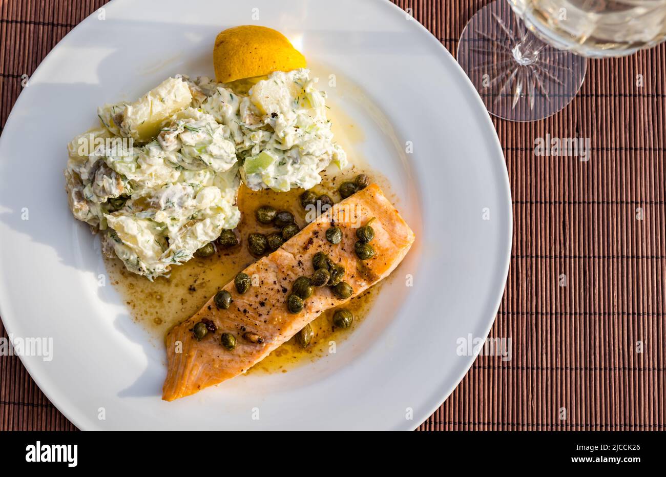 Dinner plate with slamon fillet & capers with potato salad & crystal wine glass for dinner on outdoor patio table in sunshine Stock Photo