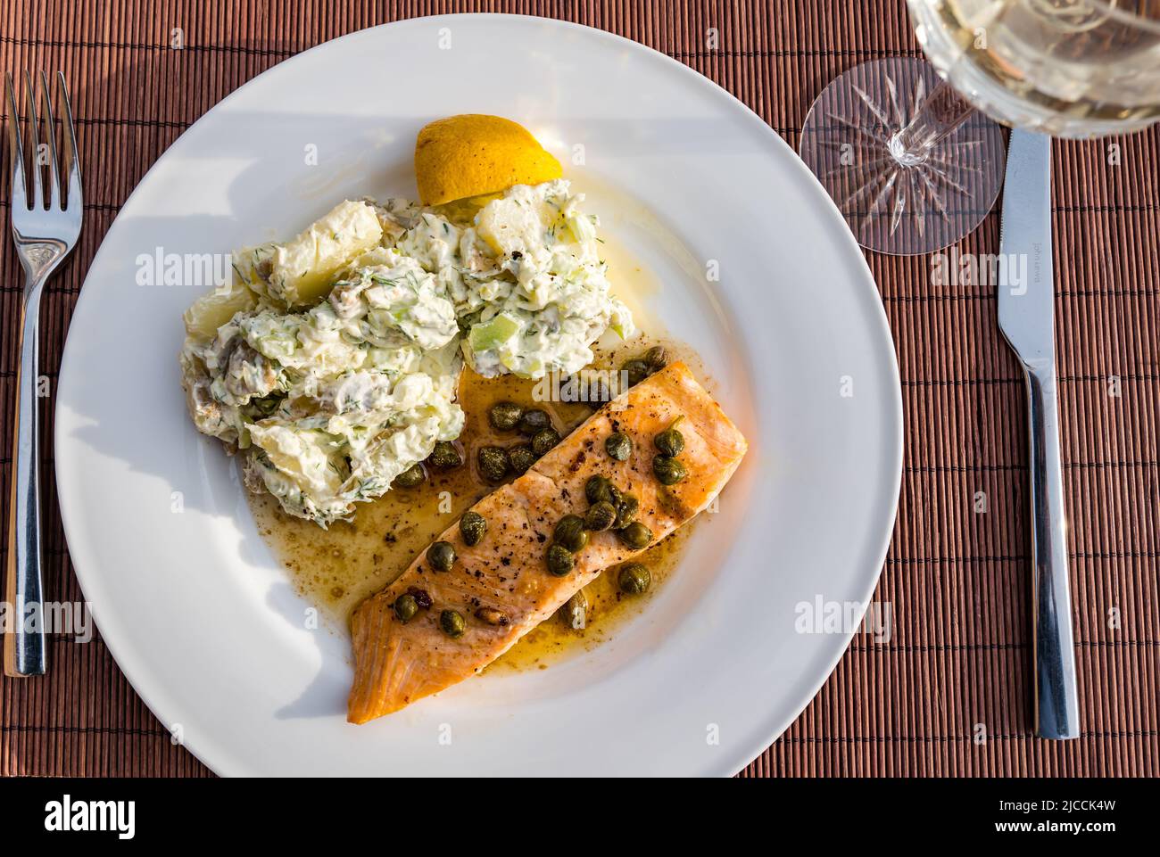 Dinner plate with slamon fillet & capers with potato salad & crystal wine glass for dinner on outdoor patio table in sunshine Stock Photo