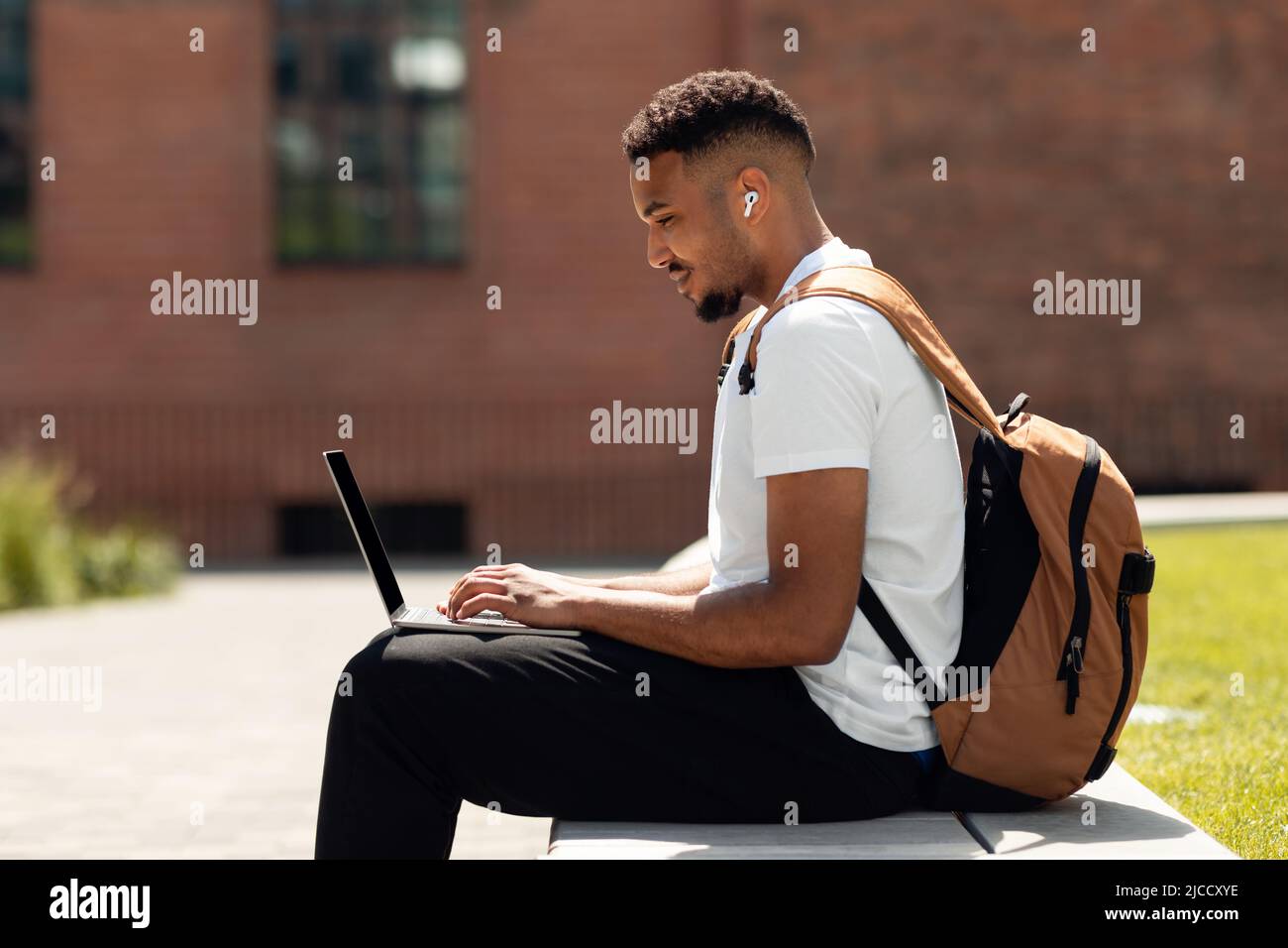 African american freelance man sitting outdoors in park, typing on laptop while working online with computer, side view Stock Photo