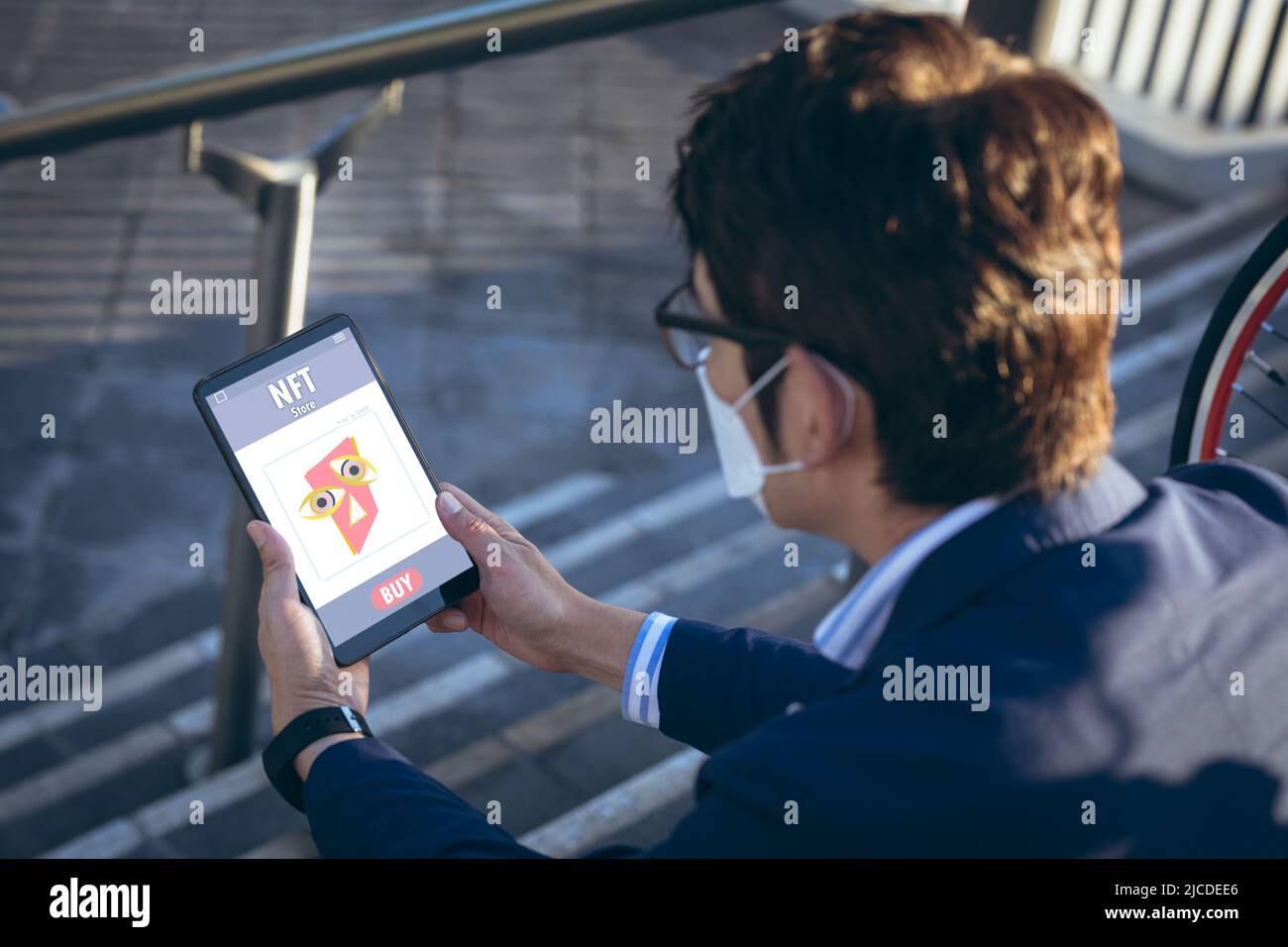 Asian businessman wearing mask doing research on nft over digital tablet while sitting on steps Stock Photo