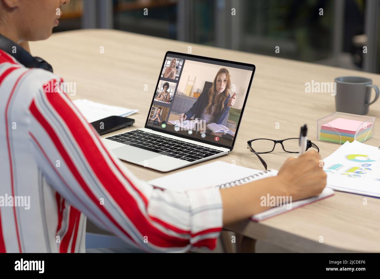 Asian businesswoman writing in diary while video conferencing with colleagues over laptop in office Stock Photo