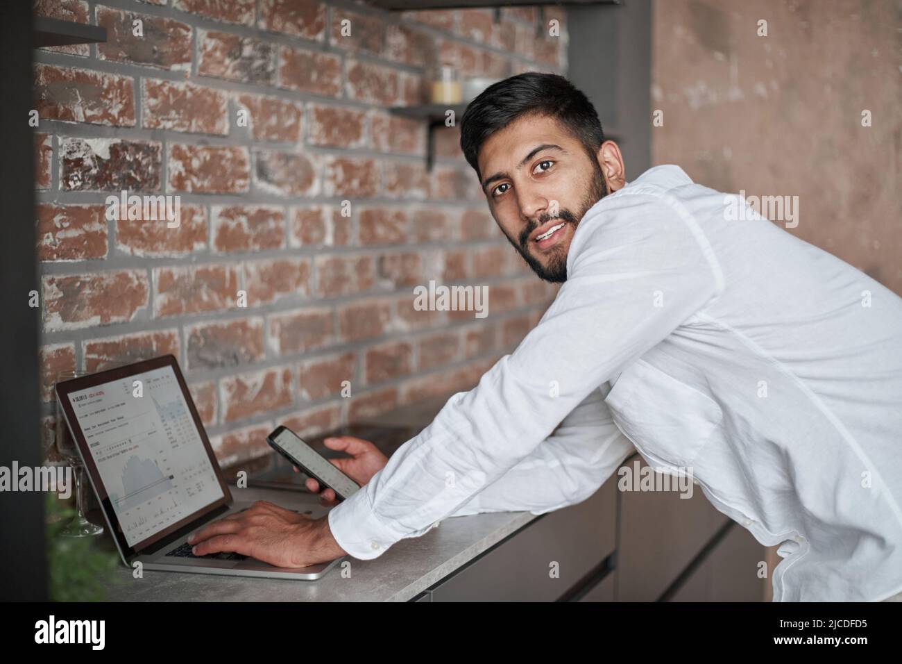 young man using a laptop and a smartphone standing in the kitchen. Stock Photo