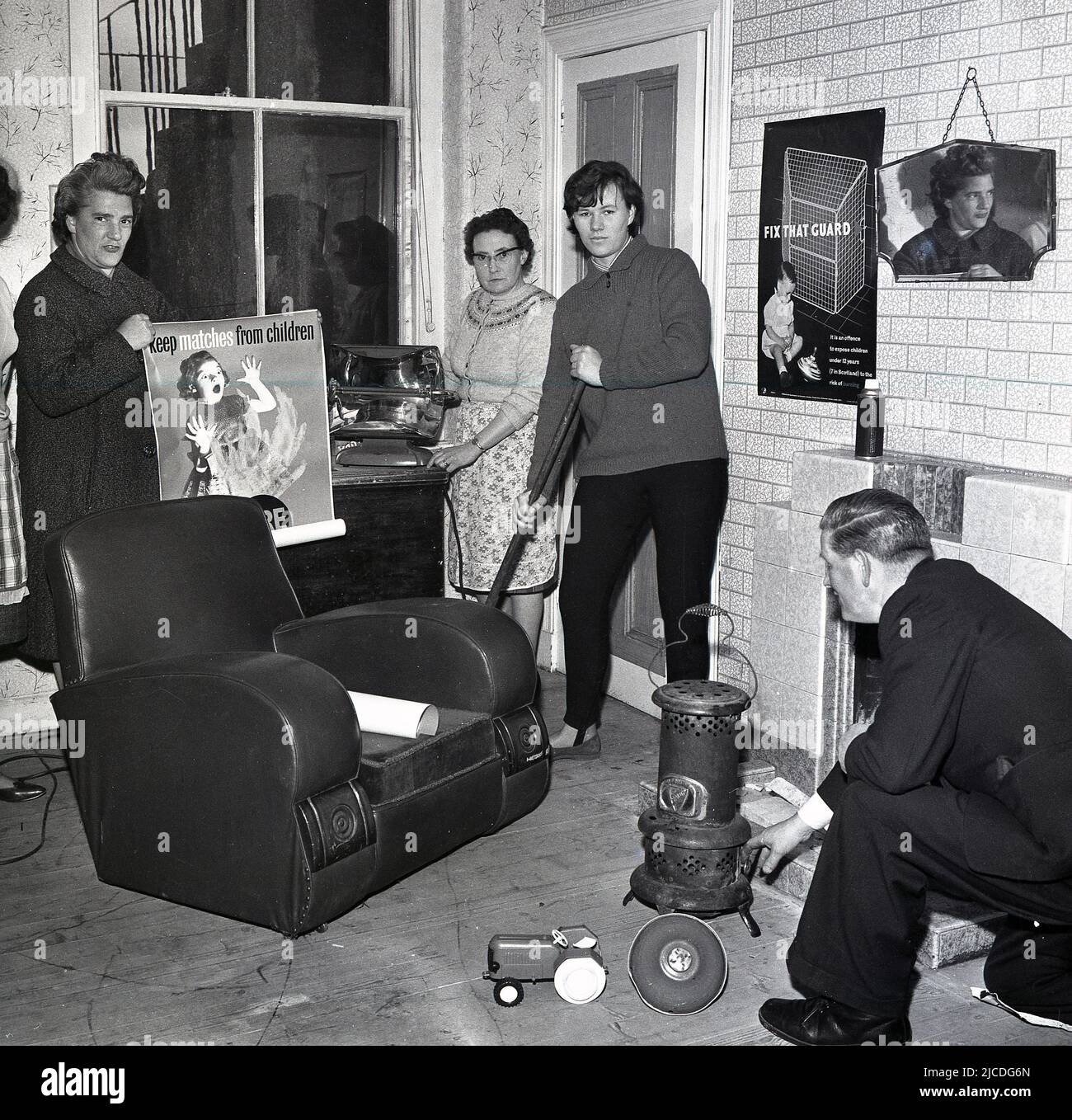 1960s, historical, fire safety education campaign, inside a room, a woman holding an electric fire and a man kneeling down by a small stove, in a picture to highlight fire prevention in the home. A lady is holding a poster warning about the danger of matches..'keep matches away from children', while another poster is on the wall highlighting the importance of fire guards...'  fix that (fire) guard', Fife, Scotland, UK. Stock Photo