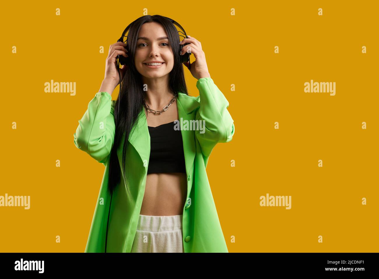Cheerful girl in green blazer wearing headphones to listen music in studio. Portrait view of happy stylish girl enjoying song, looking at camera, isolated on orange background. Concept of music fan. Stock Photo