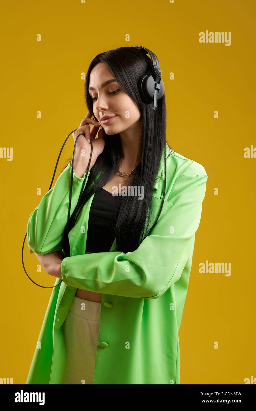 Charming long haired girl wearing trendy jacket listening music in studio. Portrait view of pretty female meloman holding wire from black earphones on head, isolated on orange. Concept of lifestyle. Stock Photo