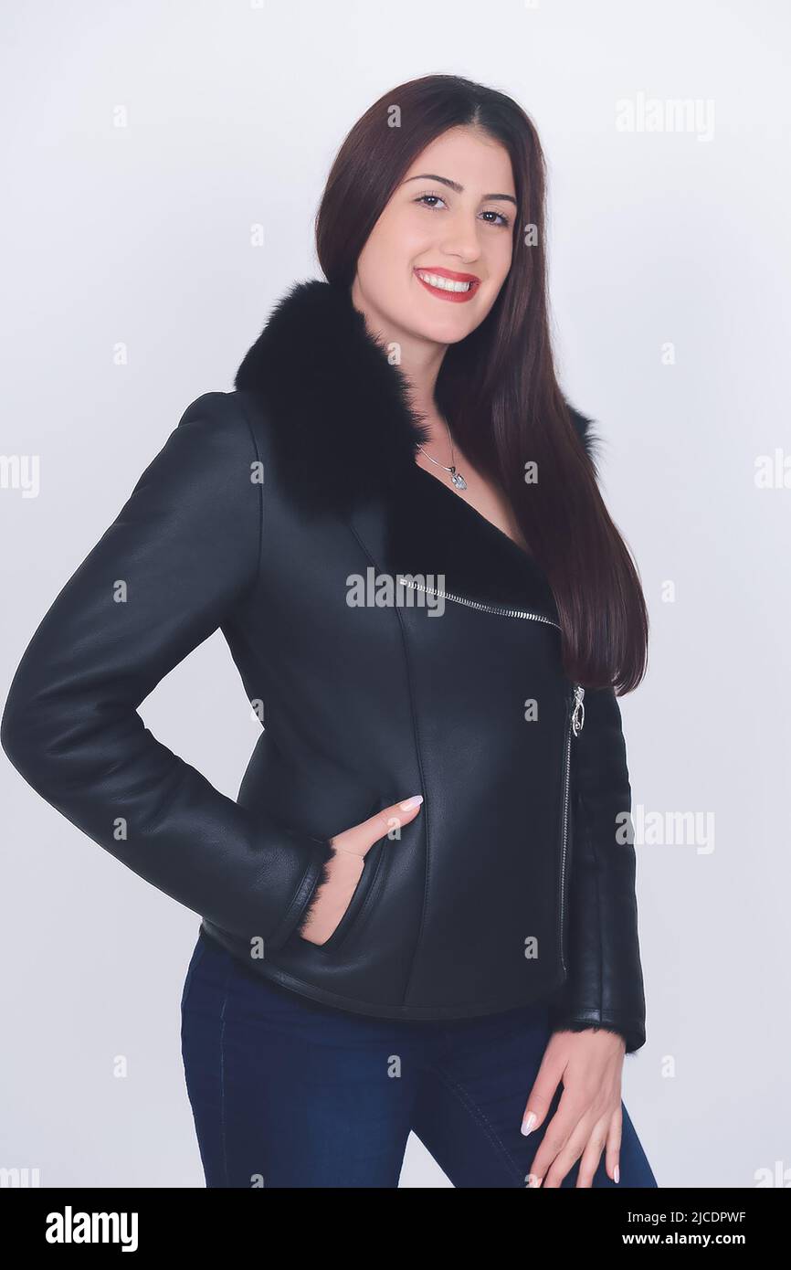 A smiling young woman in black winter leather jacket with fur on white background. Gorgeous girl in jeans looking at camera in studio. Fashion, style Stock Photo