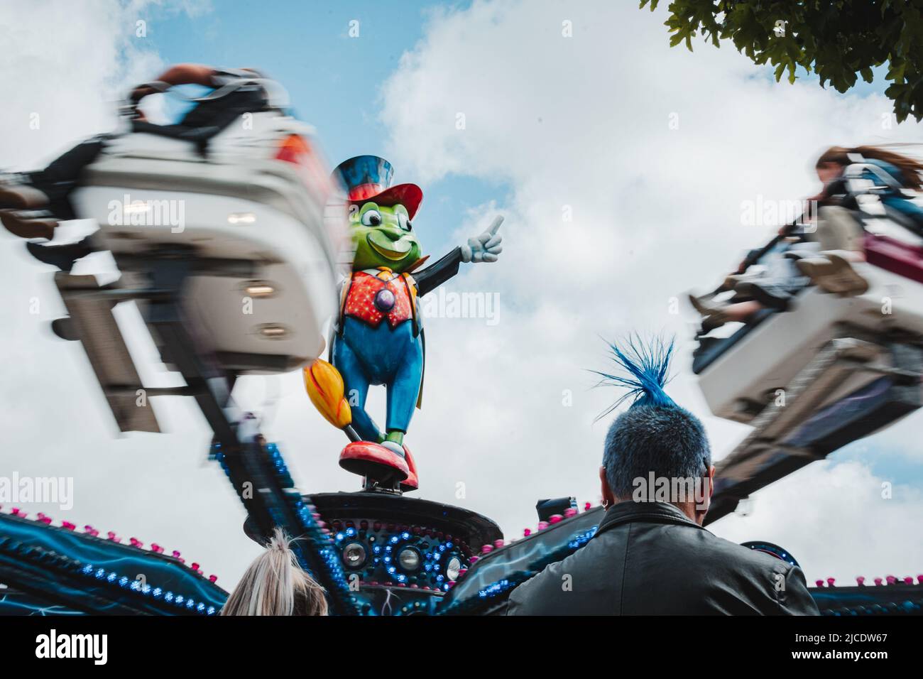 Back of a man with a Blue mohician haircut standing in front of Jiminy Cricket fair ride at Morpeth Fair Day June 2022, Northumberland Stock Photo