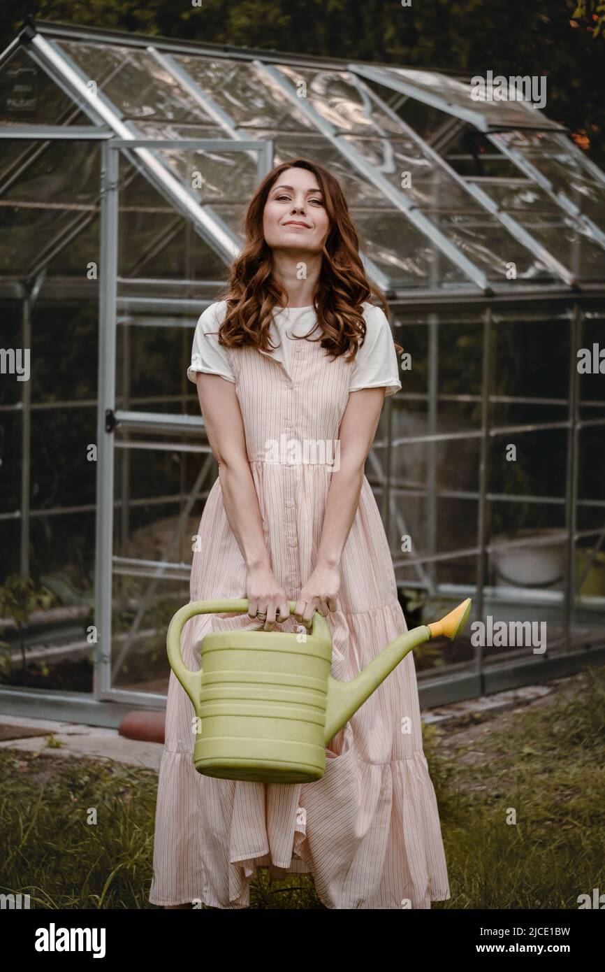 Pretty woman at her 30s in the garden with watering can walking on the grass. Beautiful dreamlike female in the evening walking in long dress over Stock Photo