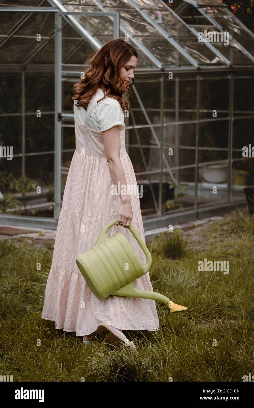 Pretty woman at her 30s in the garden with watering can walking on the grass. Beautiful dreamlike female in the evening walking in long dress over Stock Photo