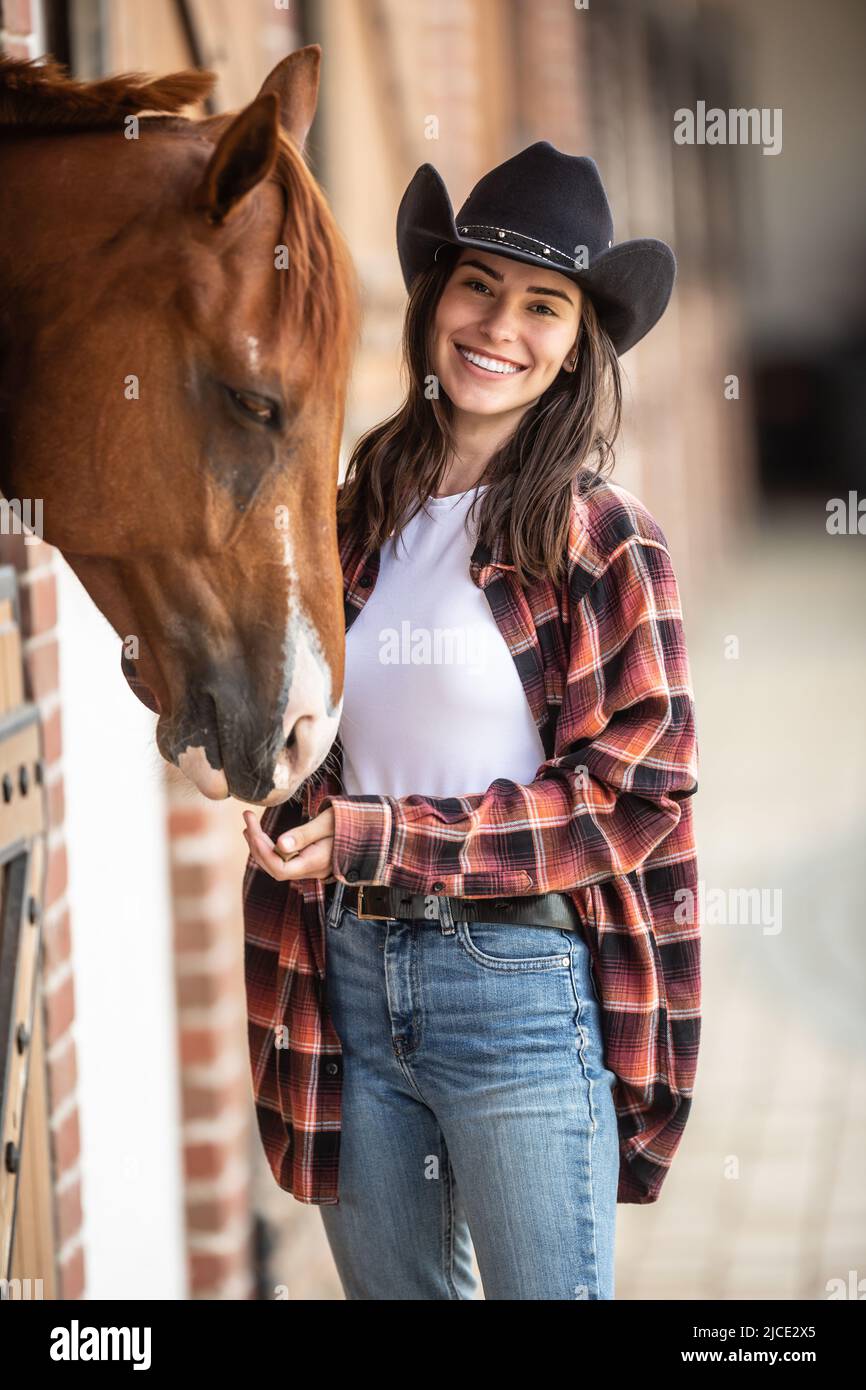 Cowgirl with a big smile feeds her horse from the hand inside the stable. Stock Photo