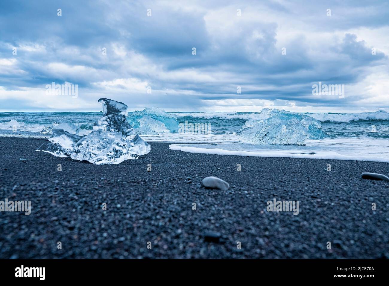 Iceberg chunks on black sand beach with waves rushing at shore against sky Stock Photo