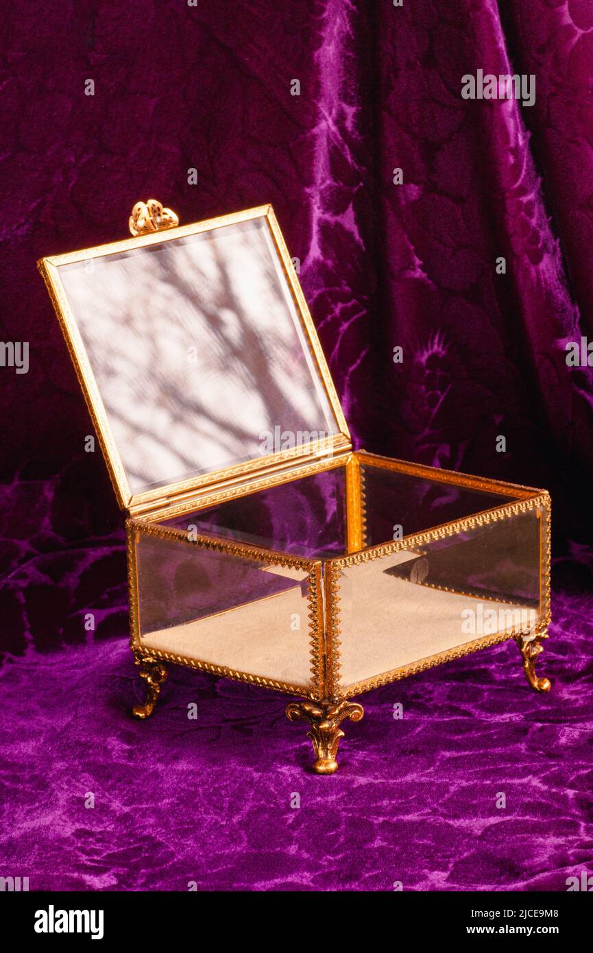 Open gold and glass filagree pedestal jewelry box with clasp on lid is empty. It sits on 4 legs with a background of draped purple velvet fabric. Stock Photo