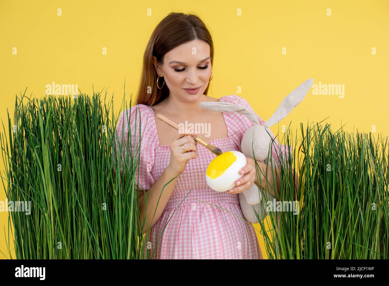 Happy woman in late pregnancy on grass decoration paint egg with brush and hold soft rabbit toy, yellow background. Portrait of young pregnant lady Stock Photo