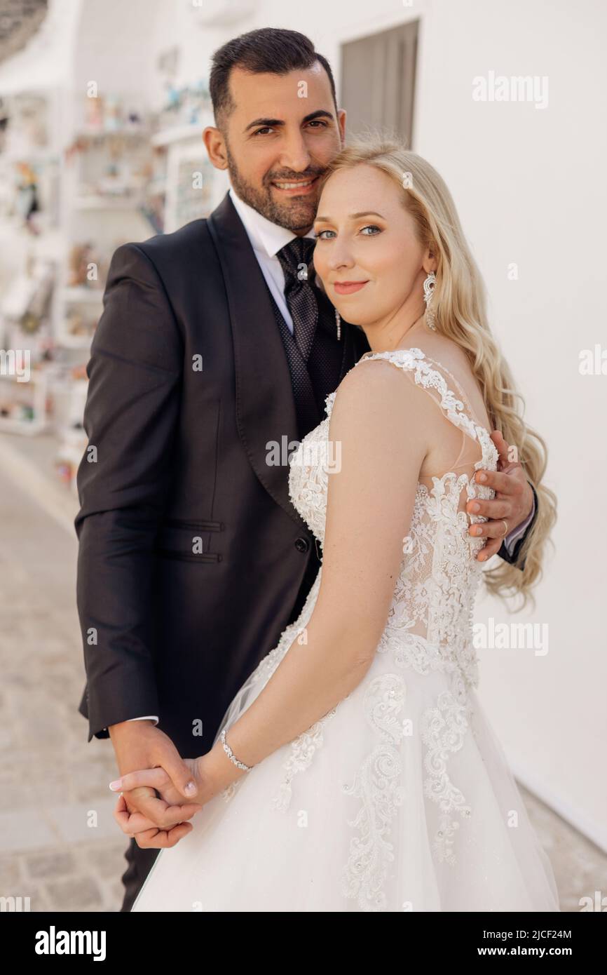 Portrait of young blonde bride in white dress and brunet groom in suit smiling and embracing while walking down street in Italy closeup, urban Stock Photo