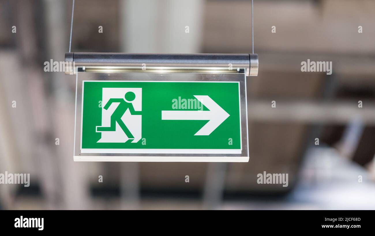 Essen, Germany - Mar 26, 2022: Illuminated emergency exit sign. With running figure and arrow to the right. Blurry background, modern design. Hanging Stock Photo