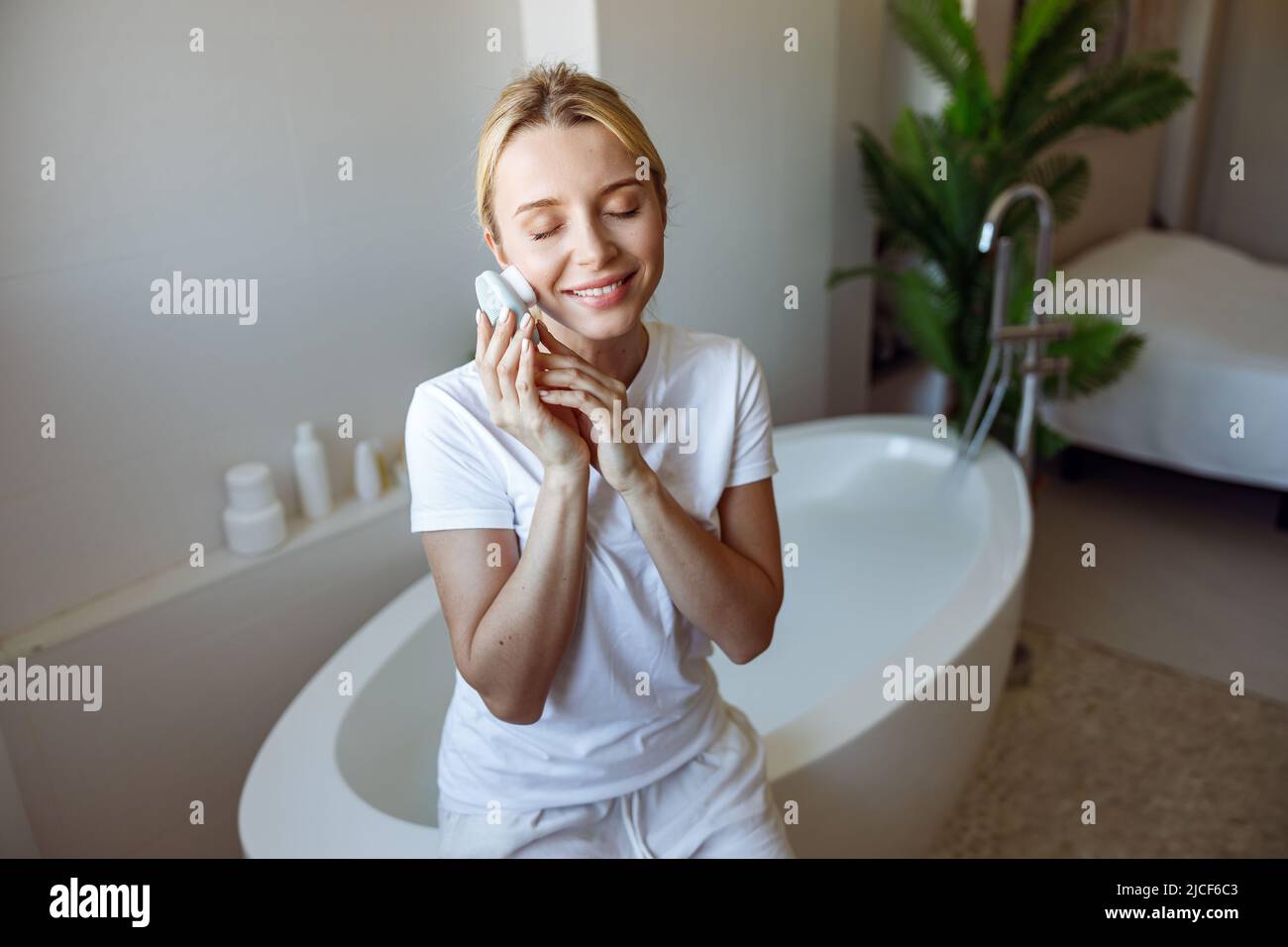 Beautiful young Caucasian woman massaging face with electro brush and smiling. At bathroom. Stock Photo