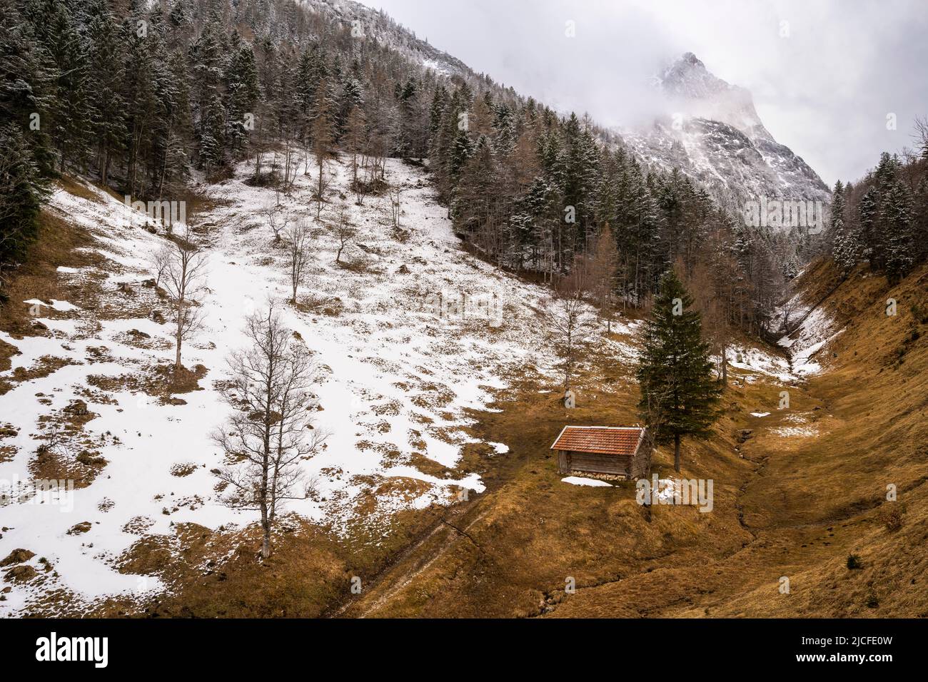A single haystack with red roof above the Ferchensee near Mittenwald in the Bavarian Alps in spring. A still partly snow-covered mountain meadow, with forest and snow, in the background the massive Wetterstein mountains with the Wetterstein in clouds and overcast sky. Stock Photo