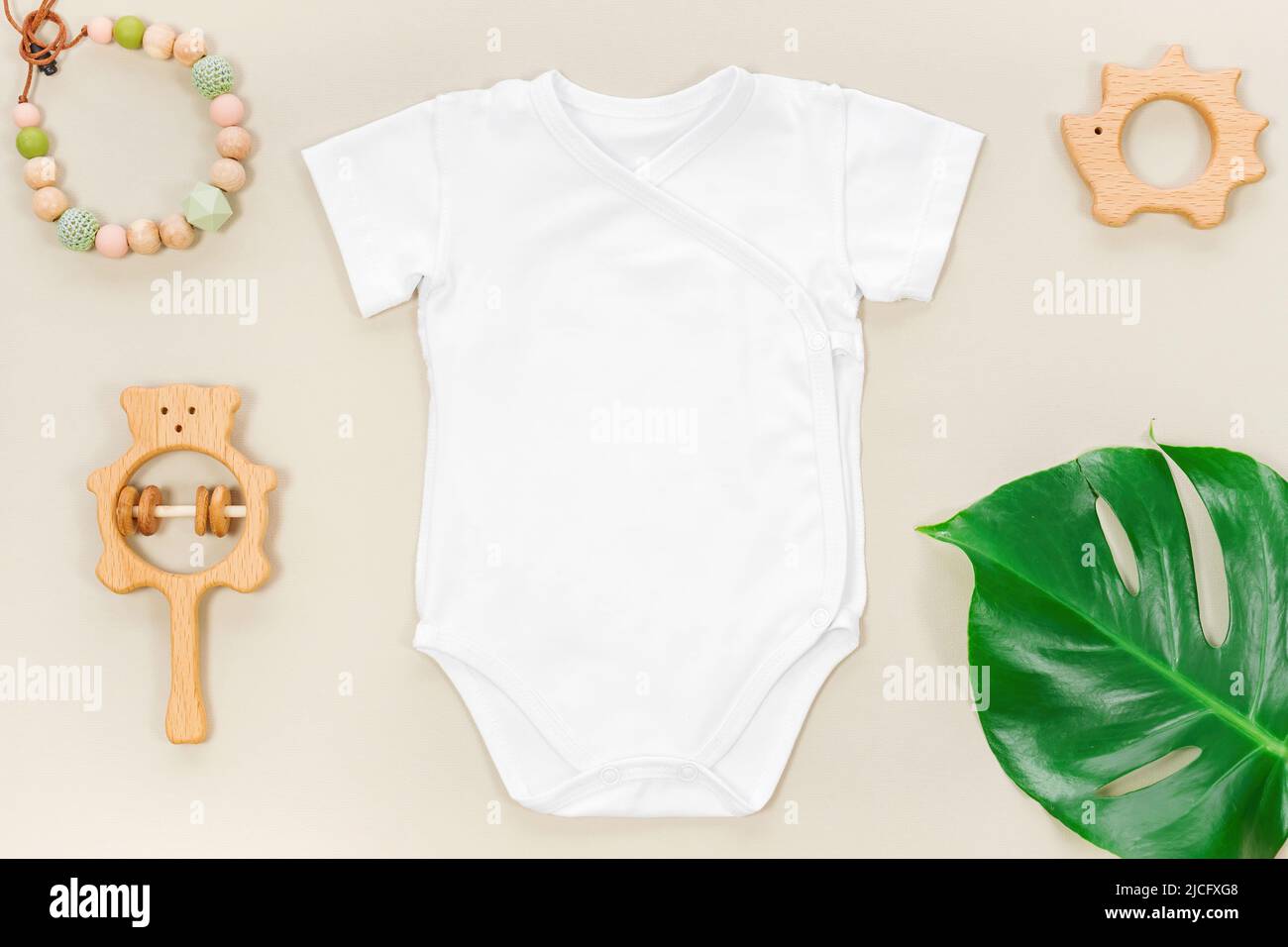 Eco friendly mockup with white wrap bodysuit and tropical monstera leaf on a pastel brown background. Teething beads and cute wooden toys for baby clo Stock Photo