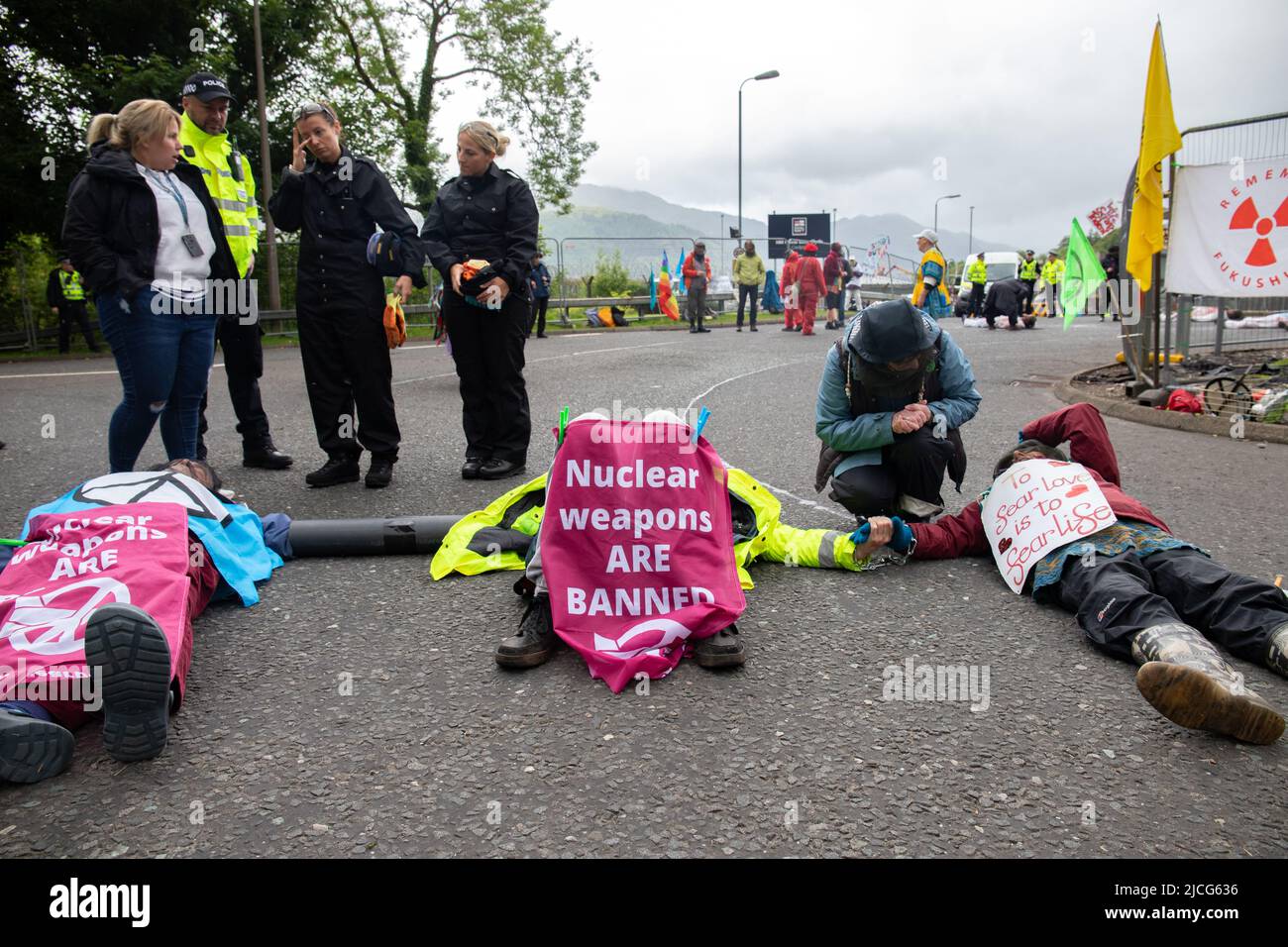 Coulport, Argyll, Scotland 13 June 2022 Five former Greenham women and three members of Peace Pirates, an offshoot of Trident Ploughshares, stage a blockade at Coulport nuclear warhead store. The ‘Greenham poured fake blood on themselves and staged a die-in in front of the gates, blocking one side of the entrance into the base while the members of ‘Peace Pirates’ (retired teacher Brian Quail, 84, with a heart transplant and disabled by a recent stroke, Willemien Hoogendorf from Holland and Gillean Lawrence, a grandmother and supporter of XR Peace) locked on blocking the roundabout further on Stock Photo