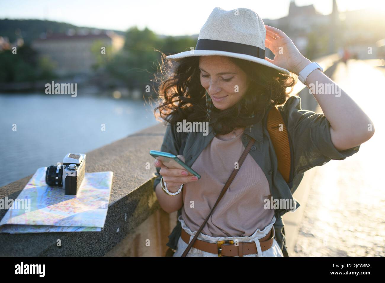 Young beautiful female traveler with map standing on bridge and using cellphone on sunny day in city. Stock Photo