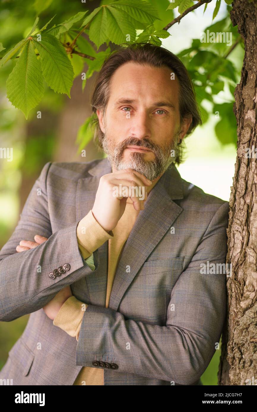 Mature handsome grey birded businessman in casual standing under tree on the grass looking at camera with arms folded touching his beard. Freelancer spend time working outdoors. Middle age crisis.  Stock Photo