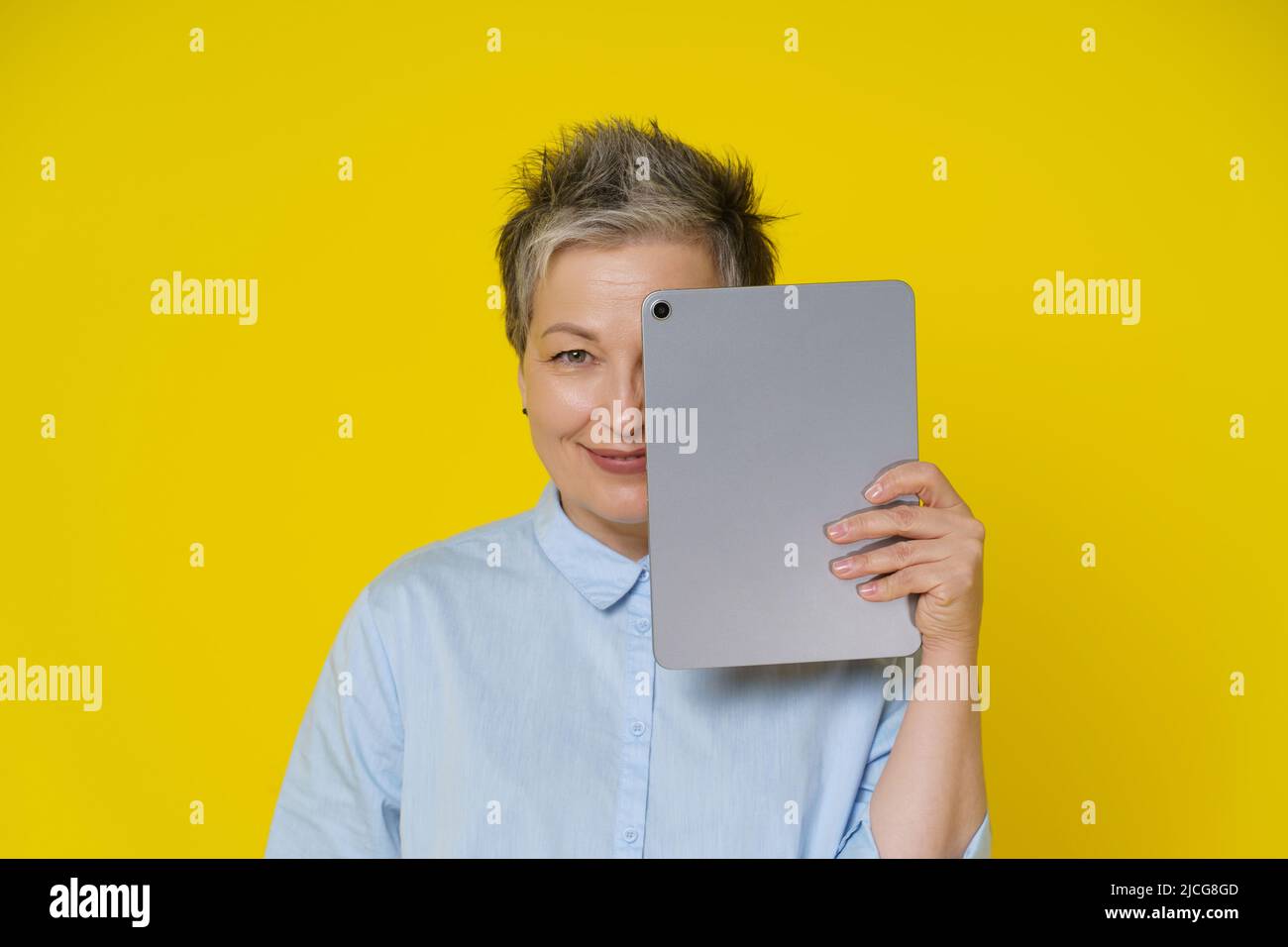 Grey haired woman hide face cover it by digital tablet smiling shy working or shopping online or checking on social media. Pretty woman in blue shirt isolated on yellow background. Stock Photo