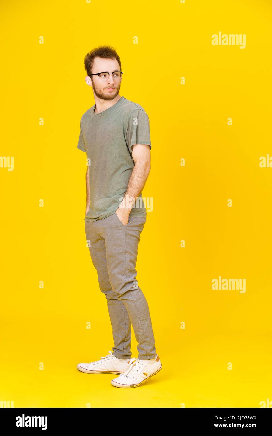 Young man wears eye glasses in casual wear posing on yellow background with hands in pockets looking sideways. Stylish bearded smart casual look hipster man. Thoughtful guy on yellow background.  Stock Photo