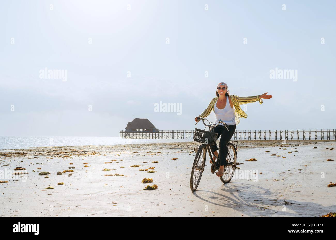 A young female dressed in light summer clothes joyfully threw up her hand while riding old vintage bicycle with a basket on the low tide ocean white s Stock Photo