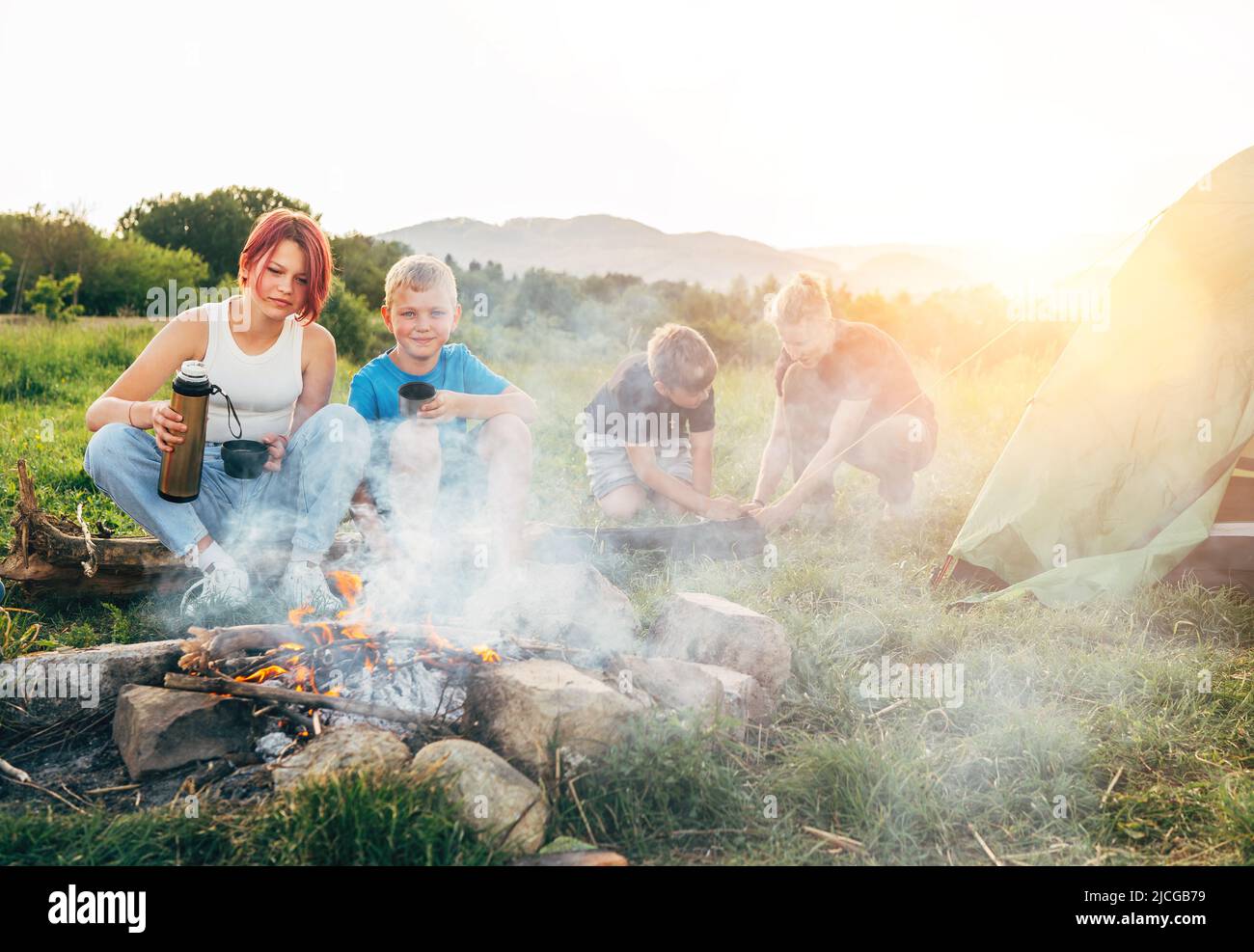 Group of smiling kids near a smoky campfire drinking tea from a thermos, two brothers set up the green tent. Happy family outdoor picnic camping activ Stock Photo