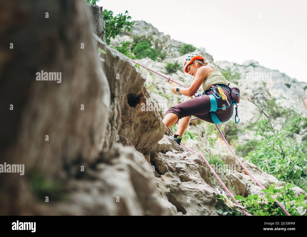 Active climber woman in protective helmet abseiling from cliff rock wall using rope with belay device and climbing harness. Active extreme sports time Stock Photo
