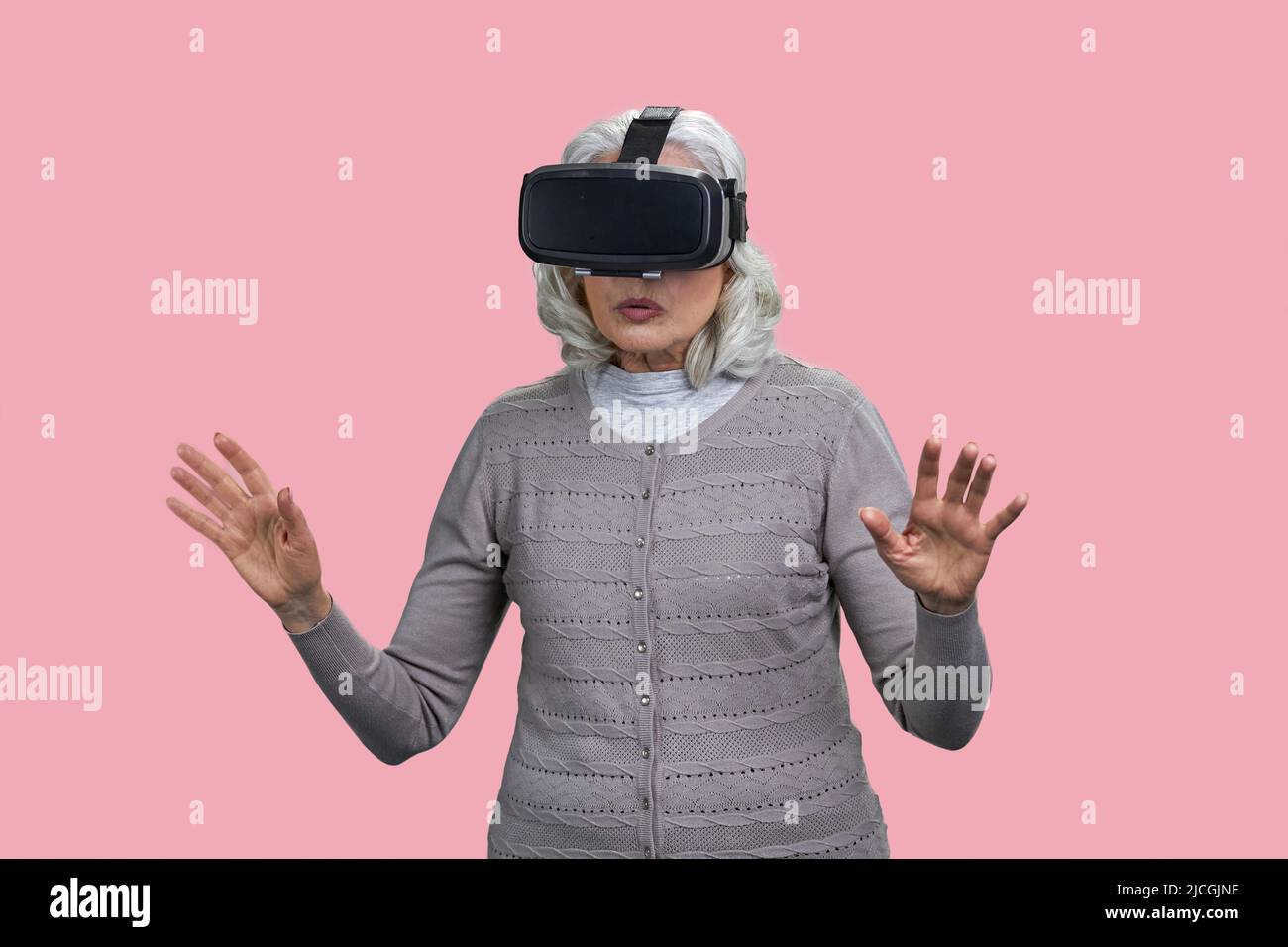 Surprised mature woman wearing virtual reality goggles standing on pink background. Seniors and modern technology concept. Stock Photo