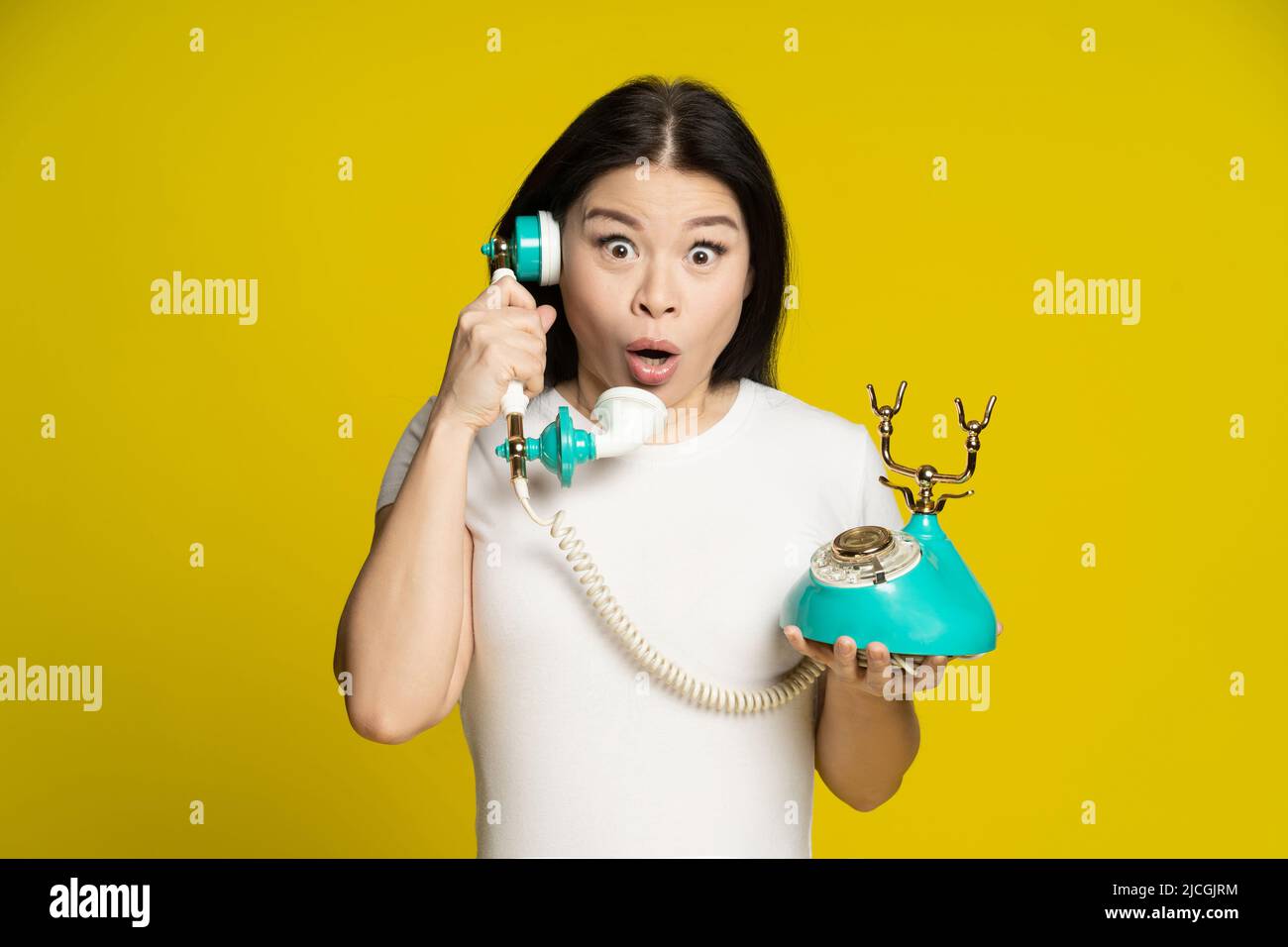 Asian woman talking using vintage, retro telephone in hands shock, excited, surprise face expression wearing white t-shirt isolated on yellow background. Communication concept.  Stock Photo