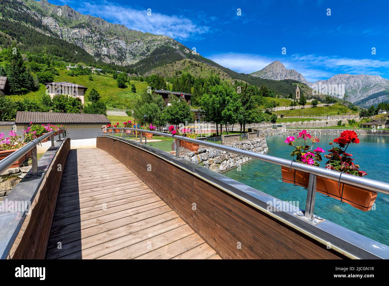 Narrow wooden bridge over small artificial lake as mountains under blue sky on background in town of Pietraporzio in Piedmont, Northern Italy. Stock Photo