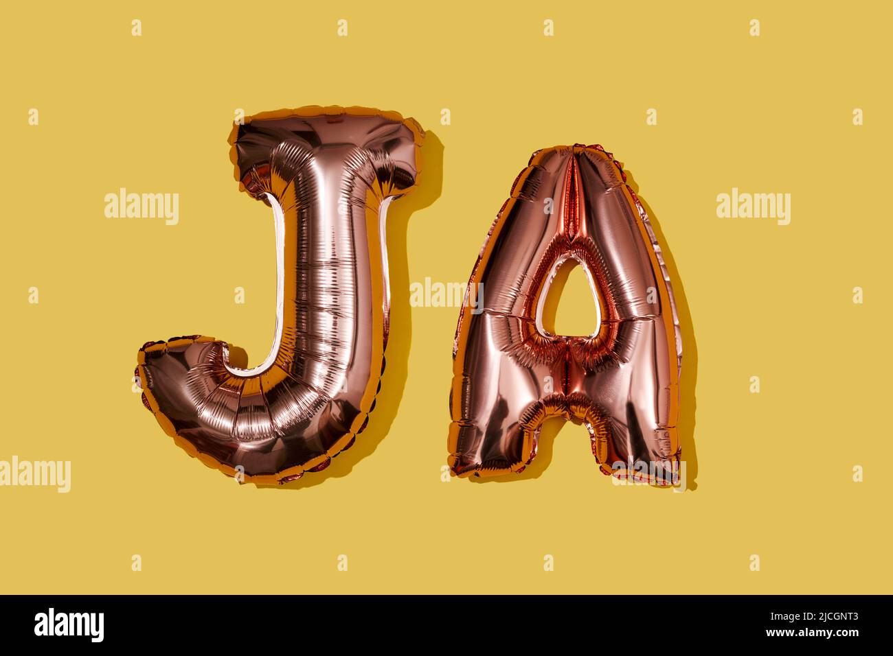some metallic pink letter-shaped balloons forming the word ja, for yes in some germanic languages, such as german, dutch or danish, on a yellow backgr Stock Photo