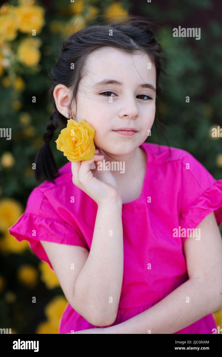 Stylish little kid girl 6-7 year old smelling yellow rose flower over blooming bushes in garden outdoor close up. Looking at camera. Springtime. Cute Stock Photo
