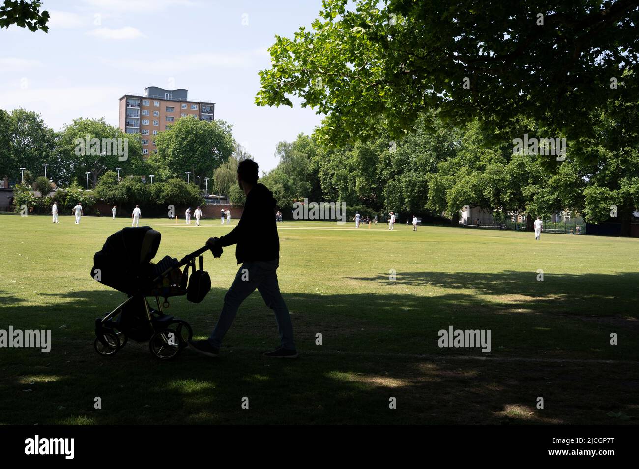 London, Hackney. London Fields. Cricket game underway with man pushing a baby in a pram. Stock Photo